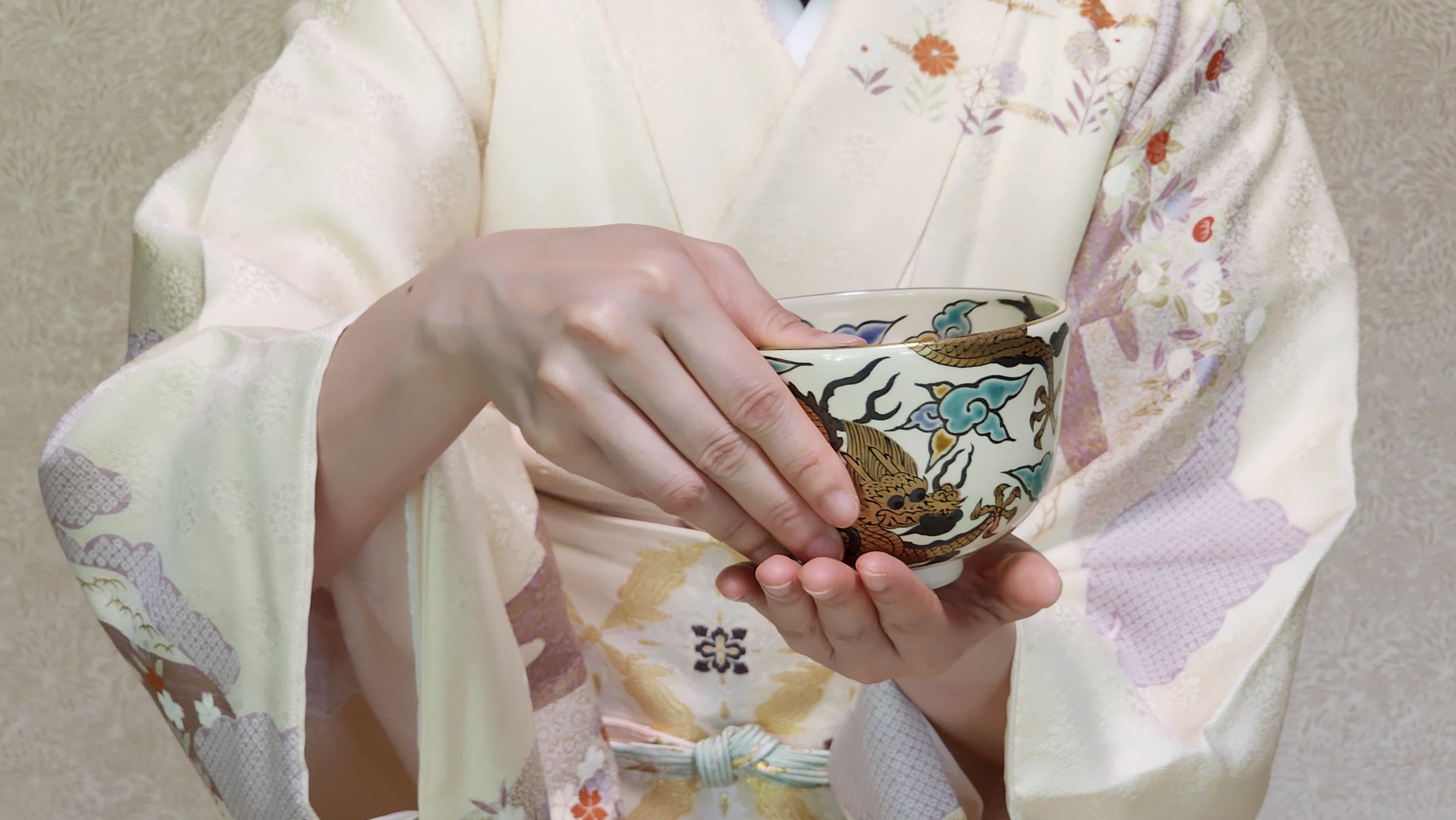 淡い色の着物をまとい、絵付けの茶碗を両手で持つ人