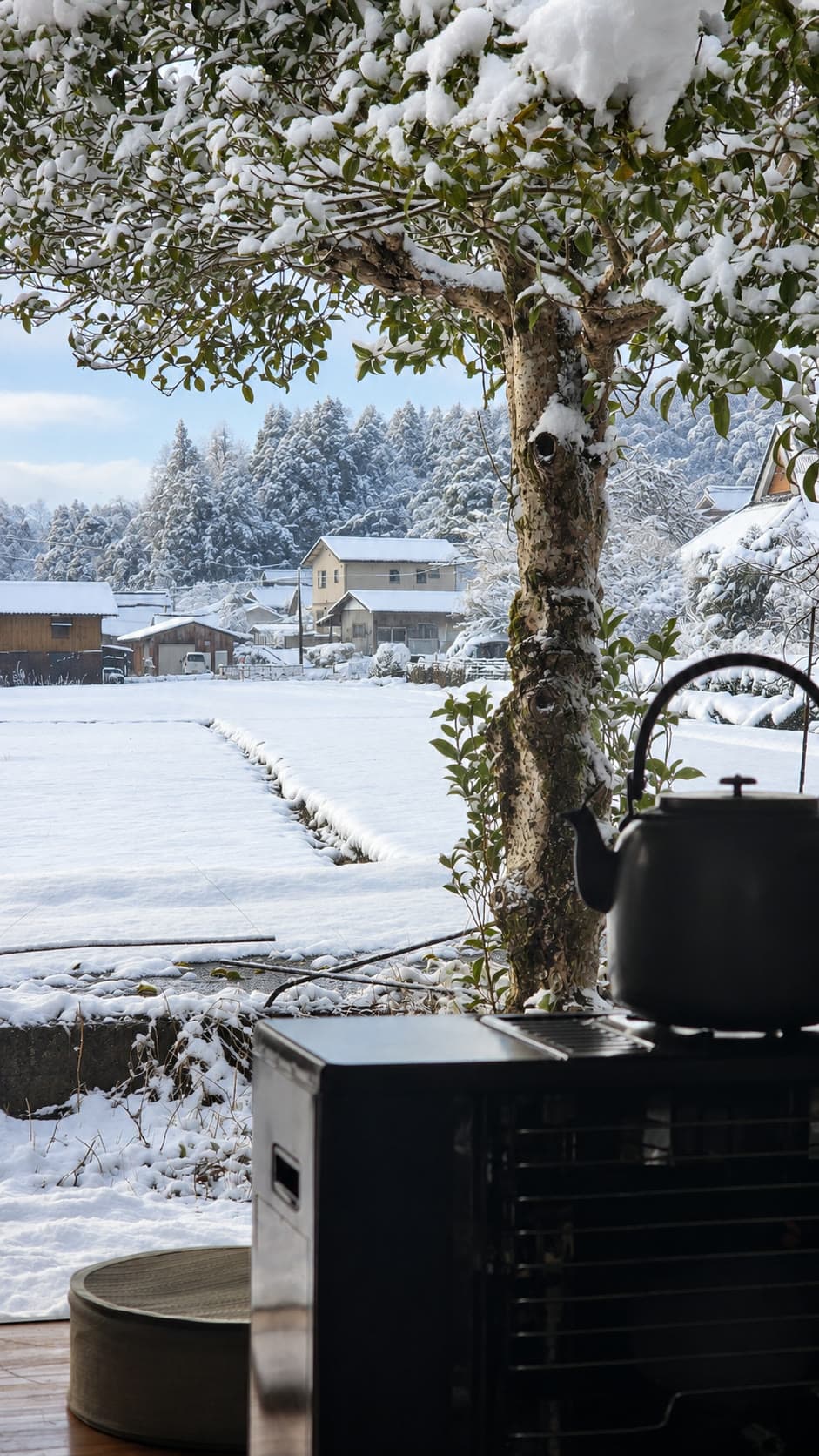 雪に包まれた山里を室内のストーブと鉄瓶越しに眺める冬景色